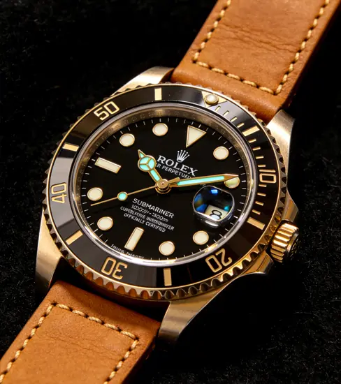 A close-up of the Coreplica diver-style watch, featuring a gold-toned and copper two-tone case with a black dial, luminescent teal-green markers, a unidirectional bezel, and a brown leather strap.